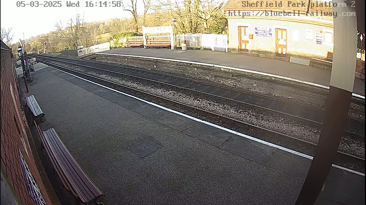 A sunny train platform with two sets of train tracks, benches, and a building in the background.