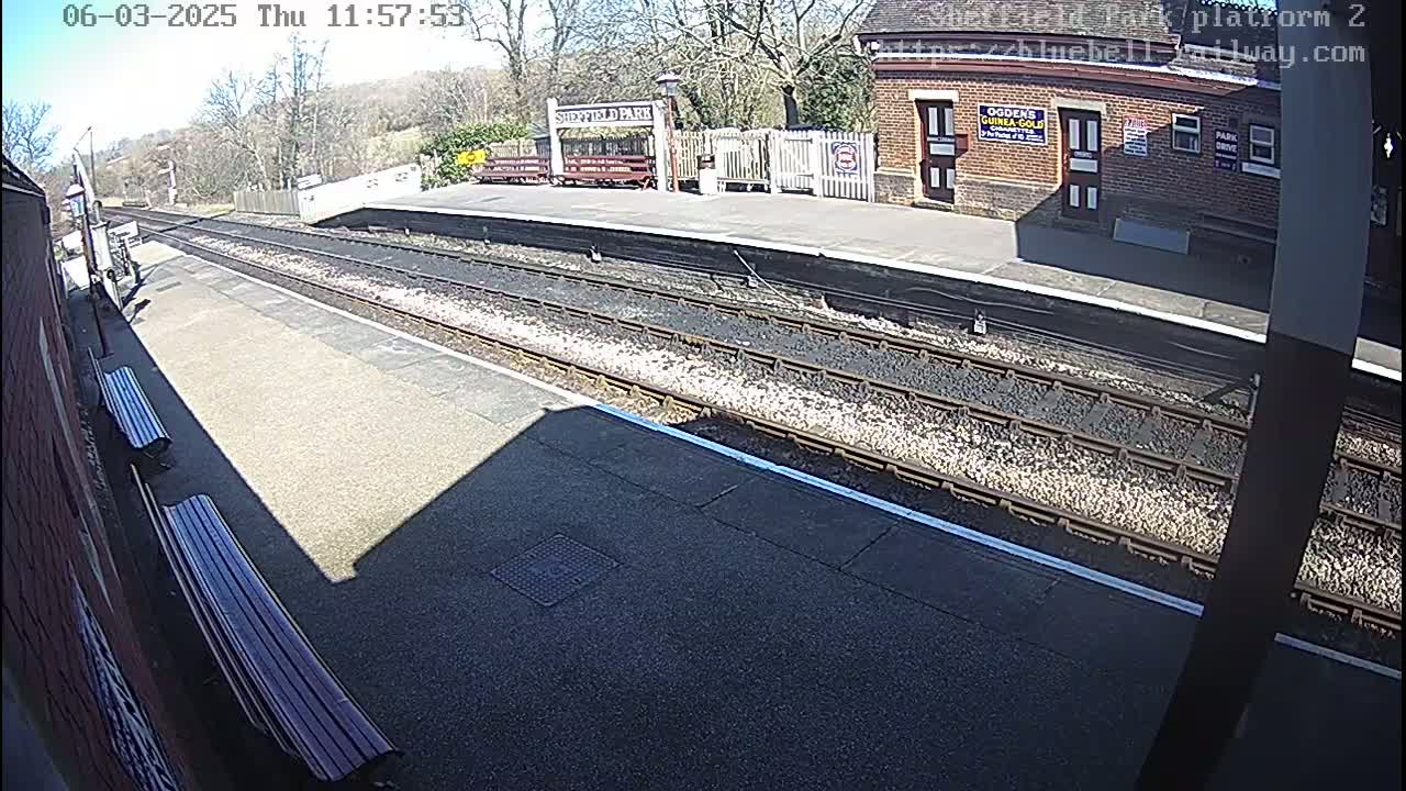 A sunny view of a train platform with two benches, train tracks, and a station building.