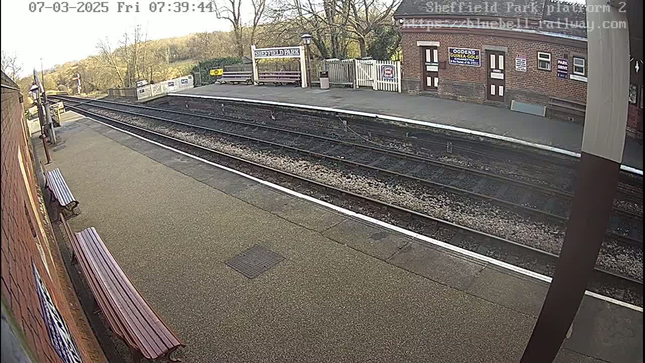 A mostly sunny view of a train platform with two benches, train tracks, and a brick station building.
