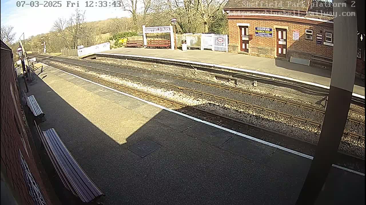 A sunny view of a train platform with two benches, train tracks, and a brick building.