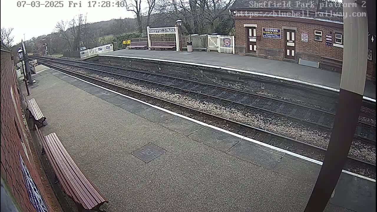 A mostly empty train platform with two parallel sets of tracks, wooden benches, and a brick station building is visible on an overcast day.