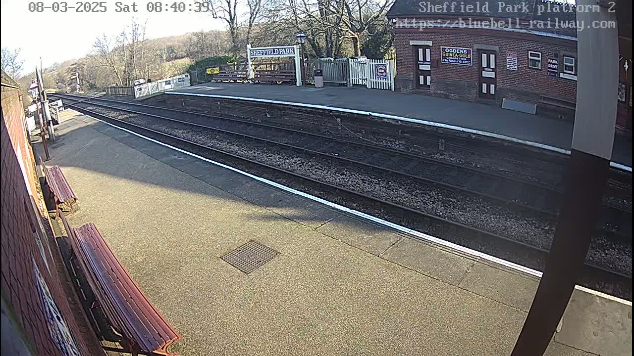 A sunny view of a mostly empty train platform with two benches and train tracks extending into the distance.