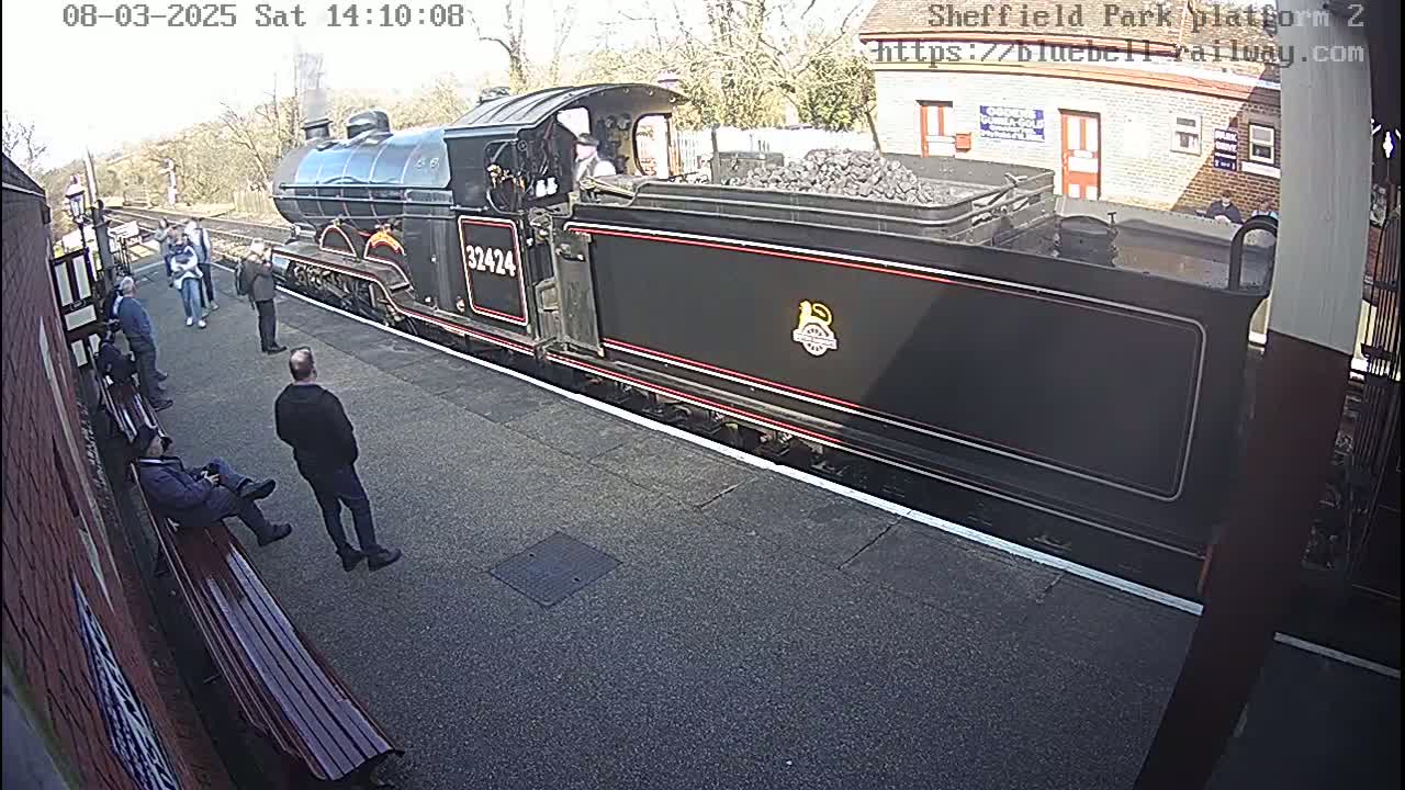 A black steam train sits at a station platform on a sunny day, with several people standing and sitting nearby.