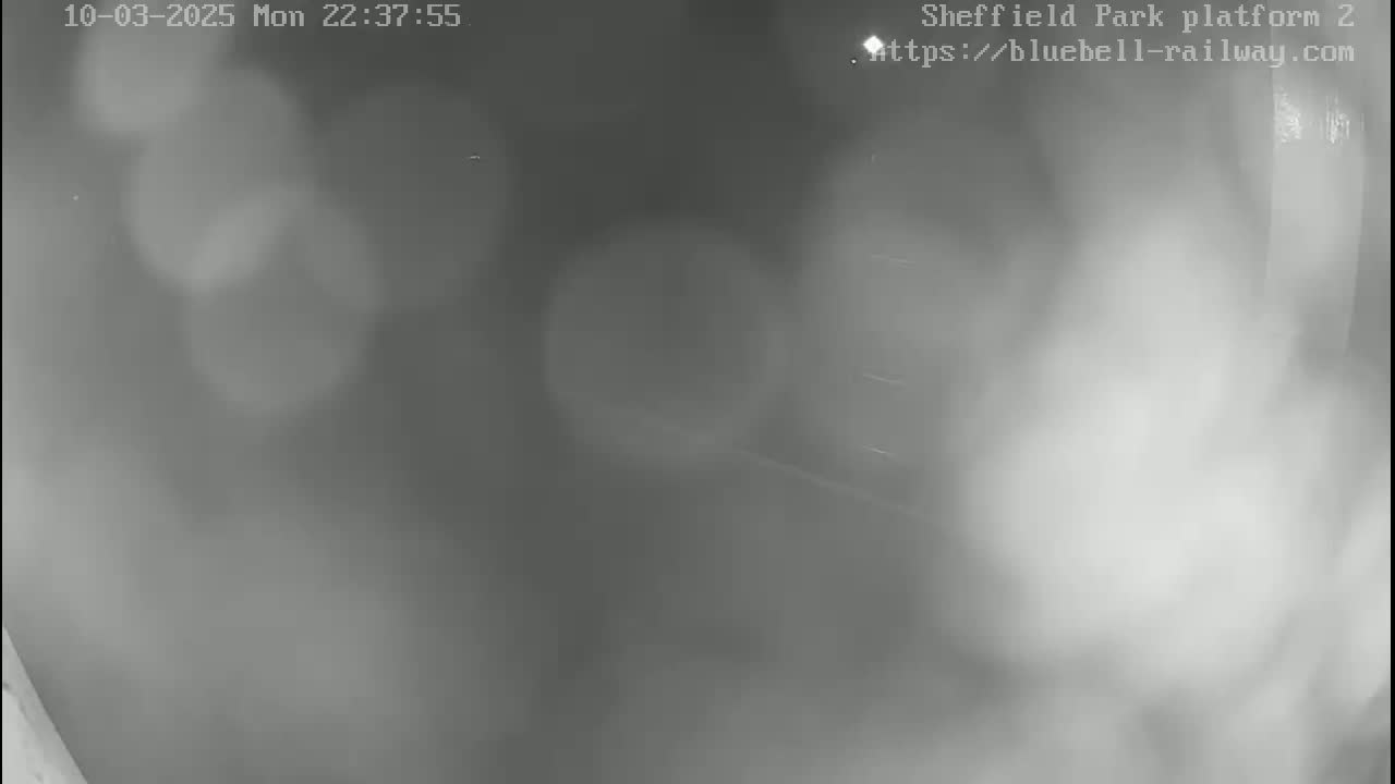A blurry, nighttime grayscale image shows a mostly empty platform with indistinct shapes and fog or mist in the air.
