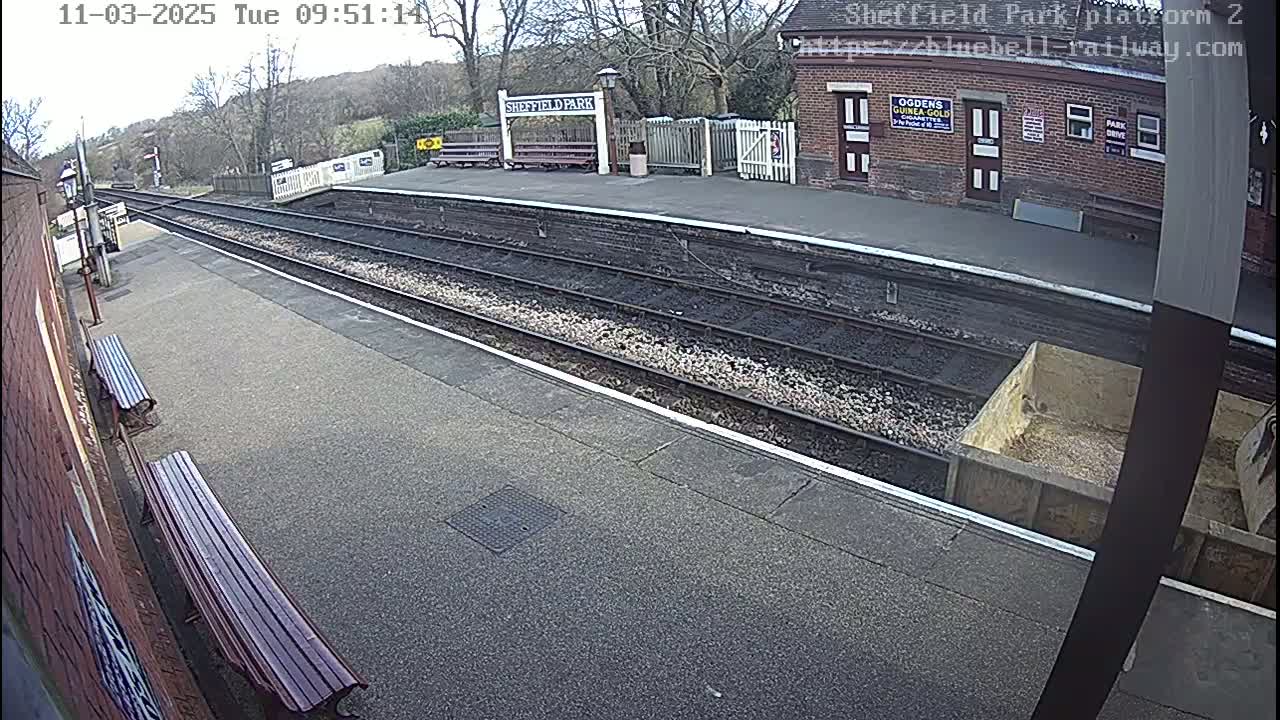 A mostly empty train platform on a partly cloudy day, with railway tracks extending into the distance and benches along the platform edge.
