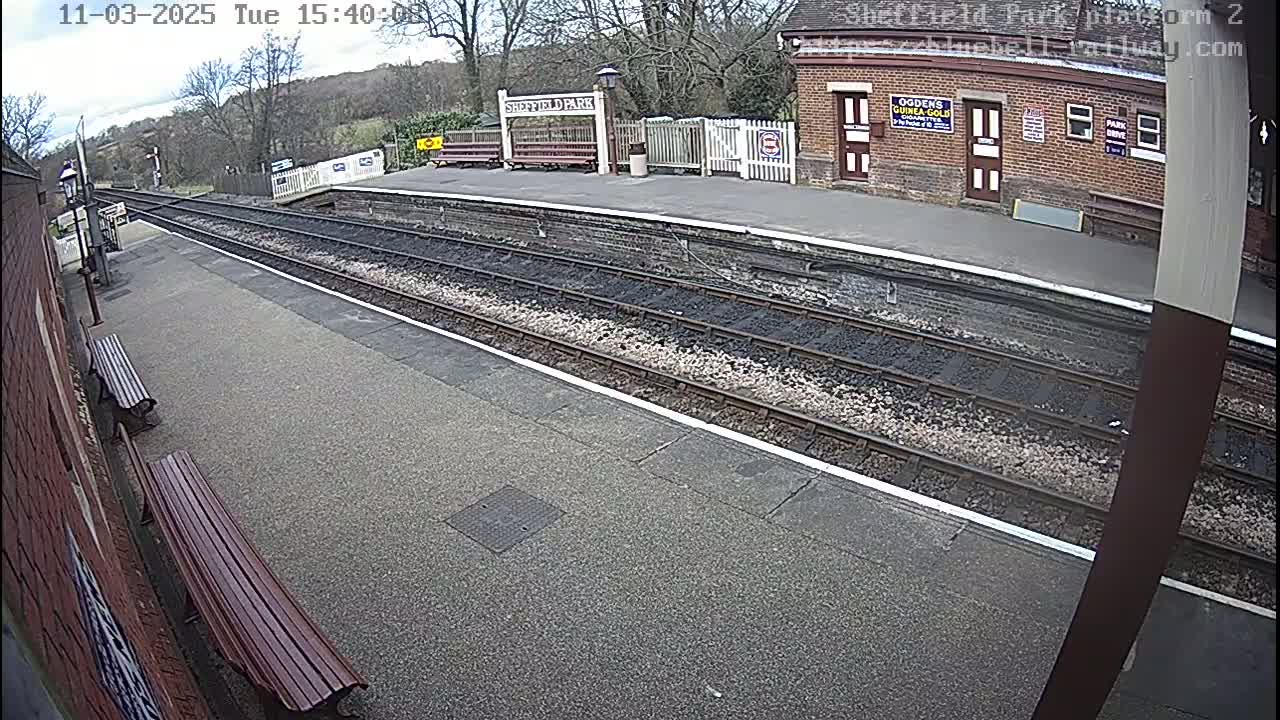 A mostly sunny view of a train platform with two benches, train tracks, and a brick building in the background.