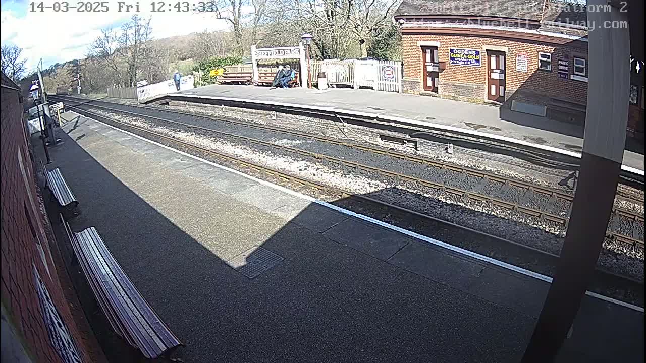 A sunny view of a train platform with two benches, a brick building, and several people standing near a shelter.