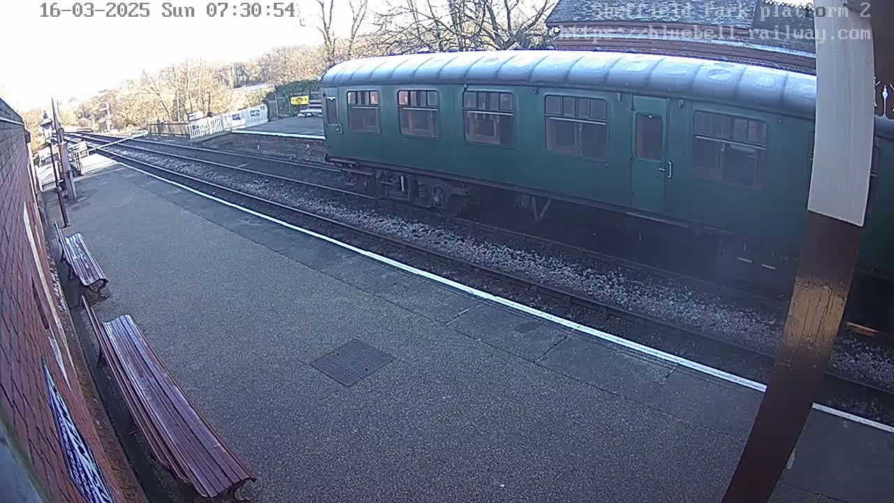 A dark-green railway carriage sits on a track at a train station on a sunny day.