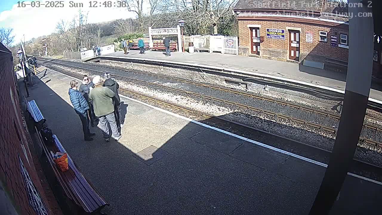 A sunny day at a train station platform shows several people standing in a group near a brick building, with train tracks and a sparsely populated platform visible.