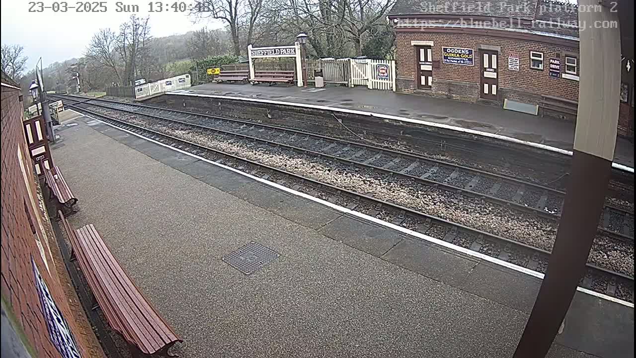 A mostly empty train platform on an overcast day with two benches and a brick station building visible.