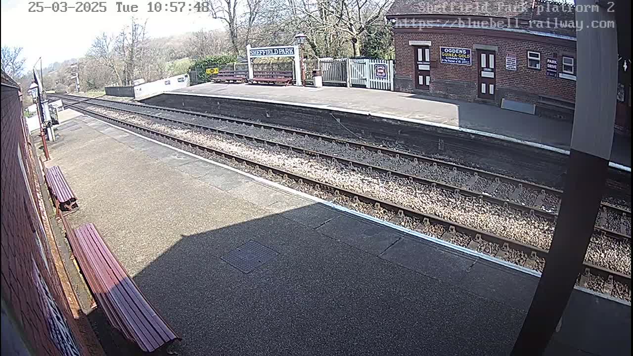 A sunny view of a train platform with two benches, train tracks, and a brick building in the background.