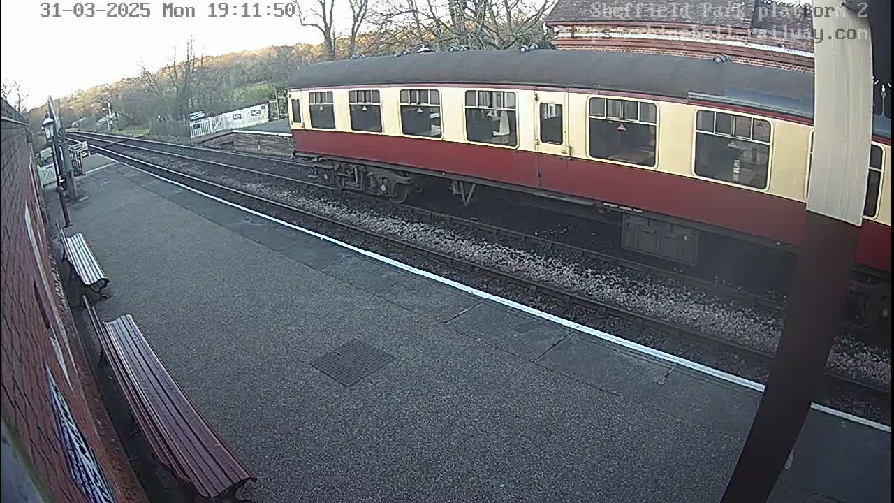 A maroon and cream passenger train car sits stationary on a train track at a station on a partly cloudy day.