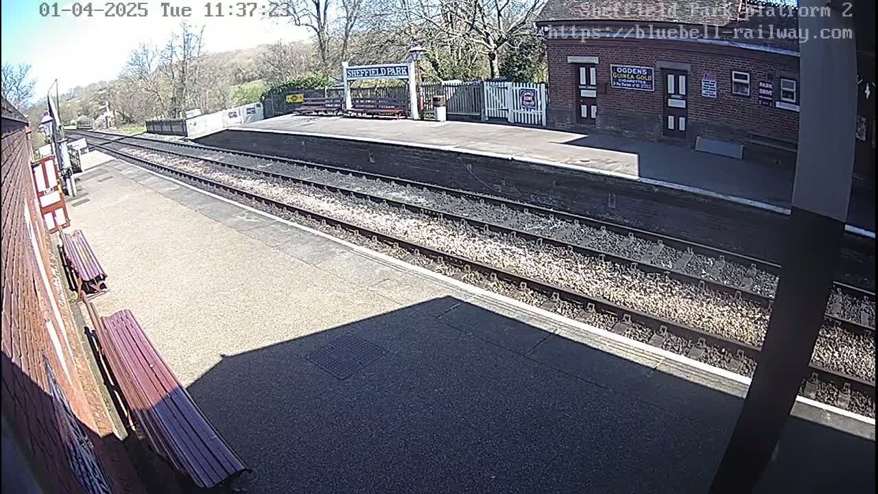 A sunny view of a train platform with two parallel train tracks extending into the distance, flanked by brick buildings and benches.