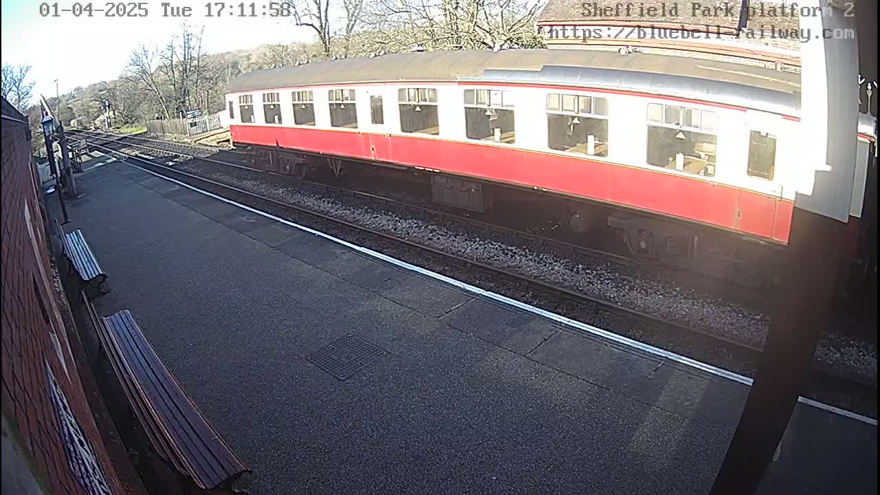 A red and white passenger train sits on a railway platform on a sunny day.