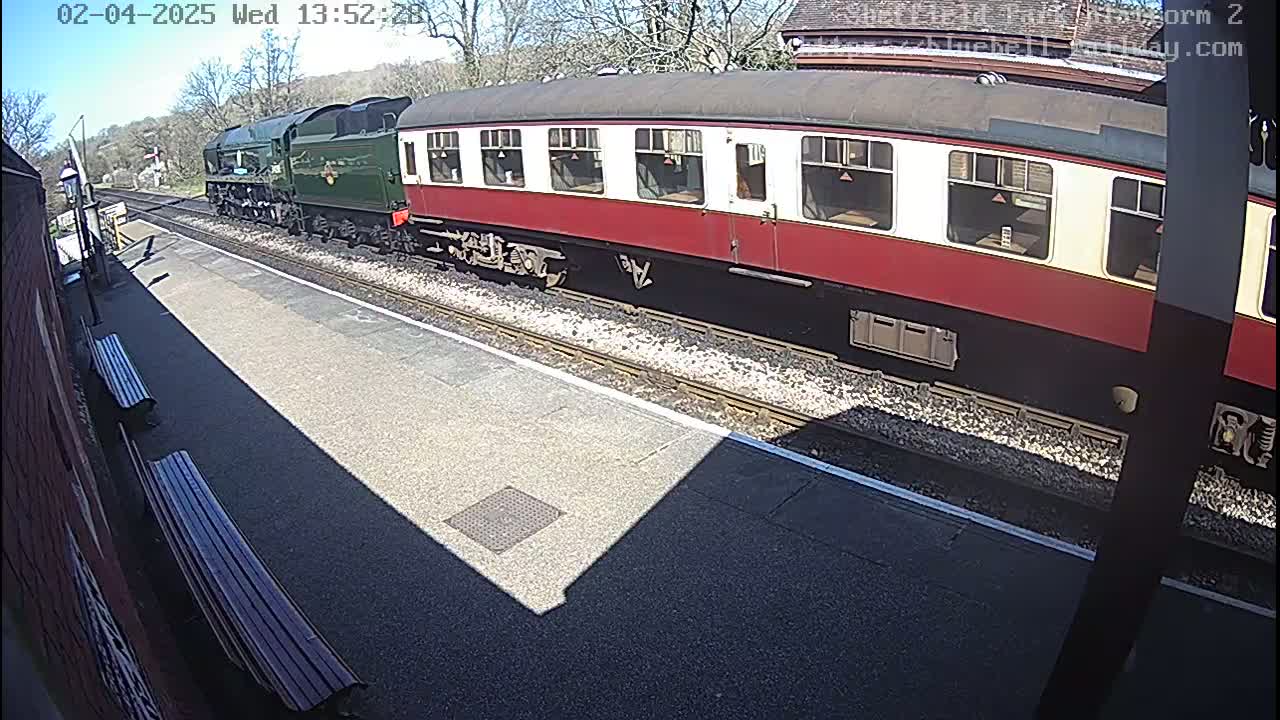 A long green and maroon passenger train sits at a station platform on a sunny day.