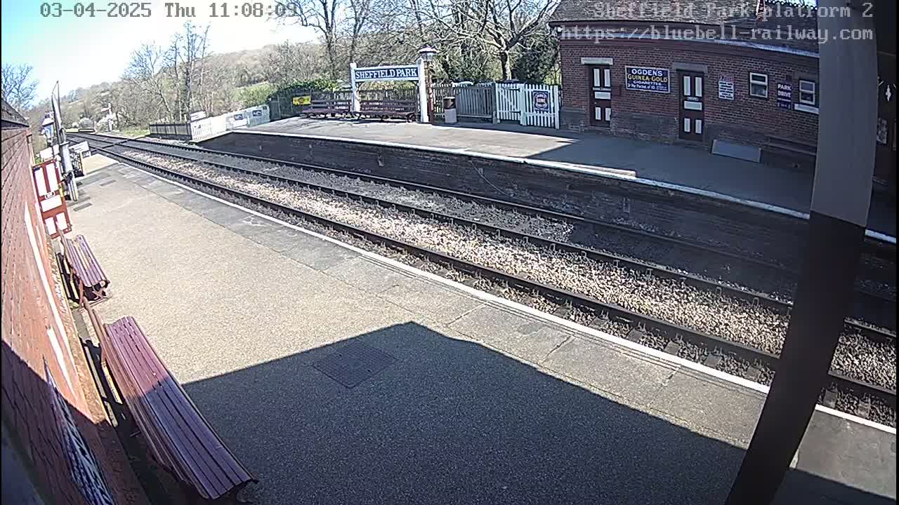 A sunny view of a train platform with two parallel sets of tracks and benches along the side of a brick building.