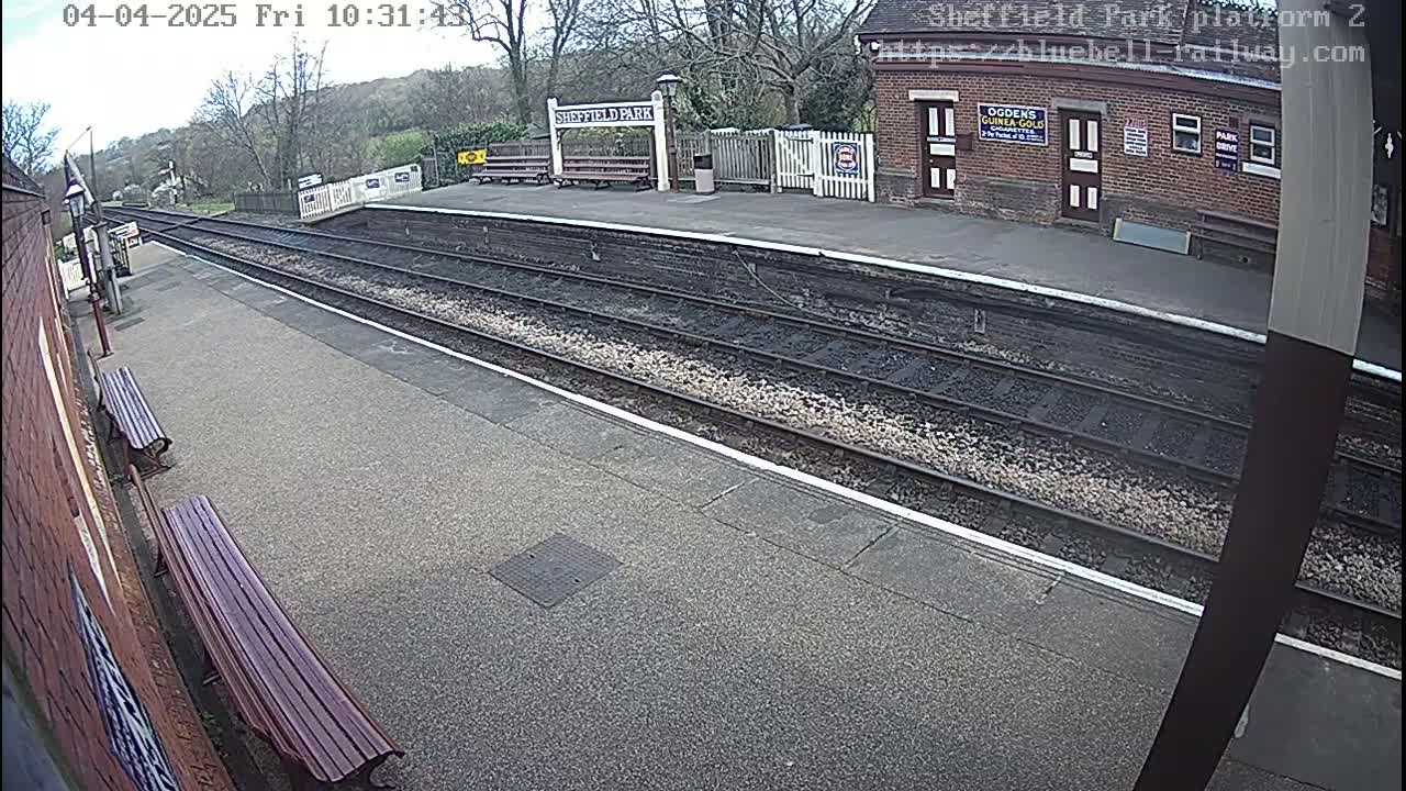 A mostly empty train platform on a cloudy day, with two benches and a brick building visible in the background.