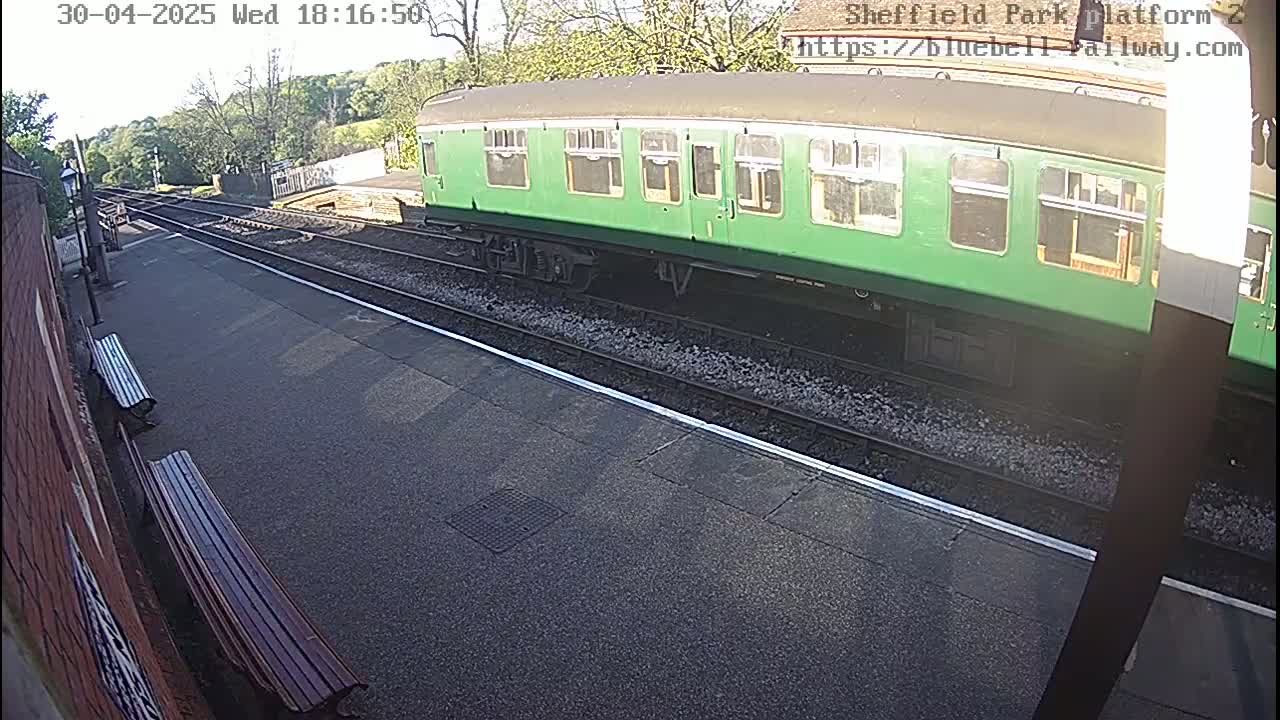 A long green train car sits on a railway track next to a platform with two benches on a sunny day.