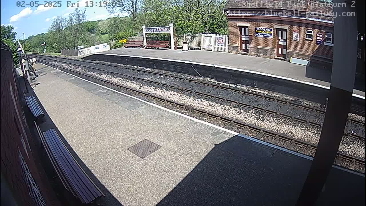 A sunny view of a train platform with two tracks, benches, and a brick building in the background.