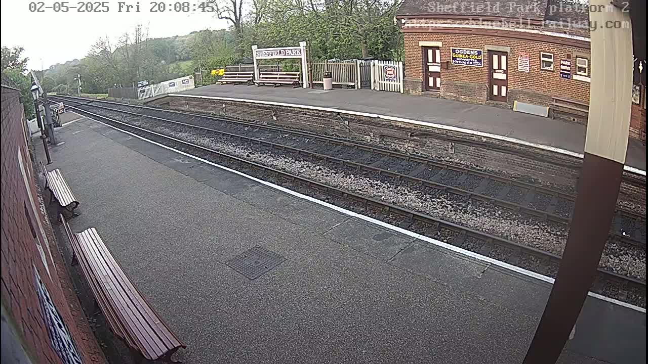 A train platform with two benches, train tracks, and a brick building is visible under an overcast sky.