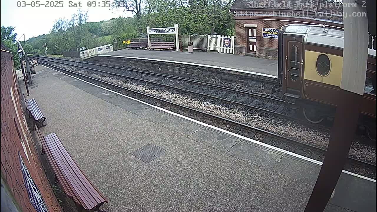 A train car is partially visible at a train station platform on a partly sunny day, with trees and greenery in the background.