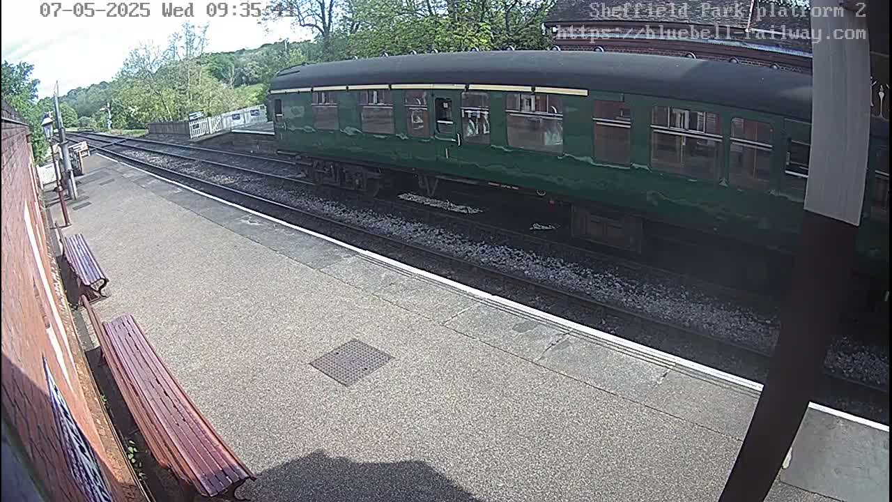 A long, dark-green passenger train sits at a station platform on a sunny day.