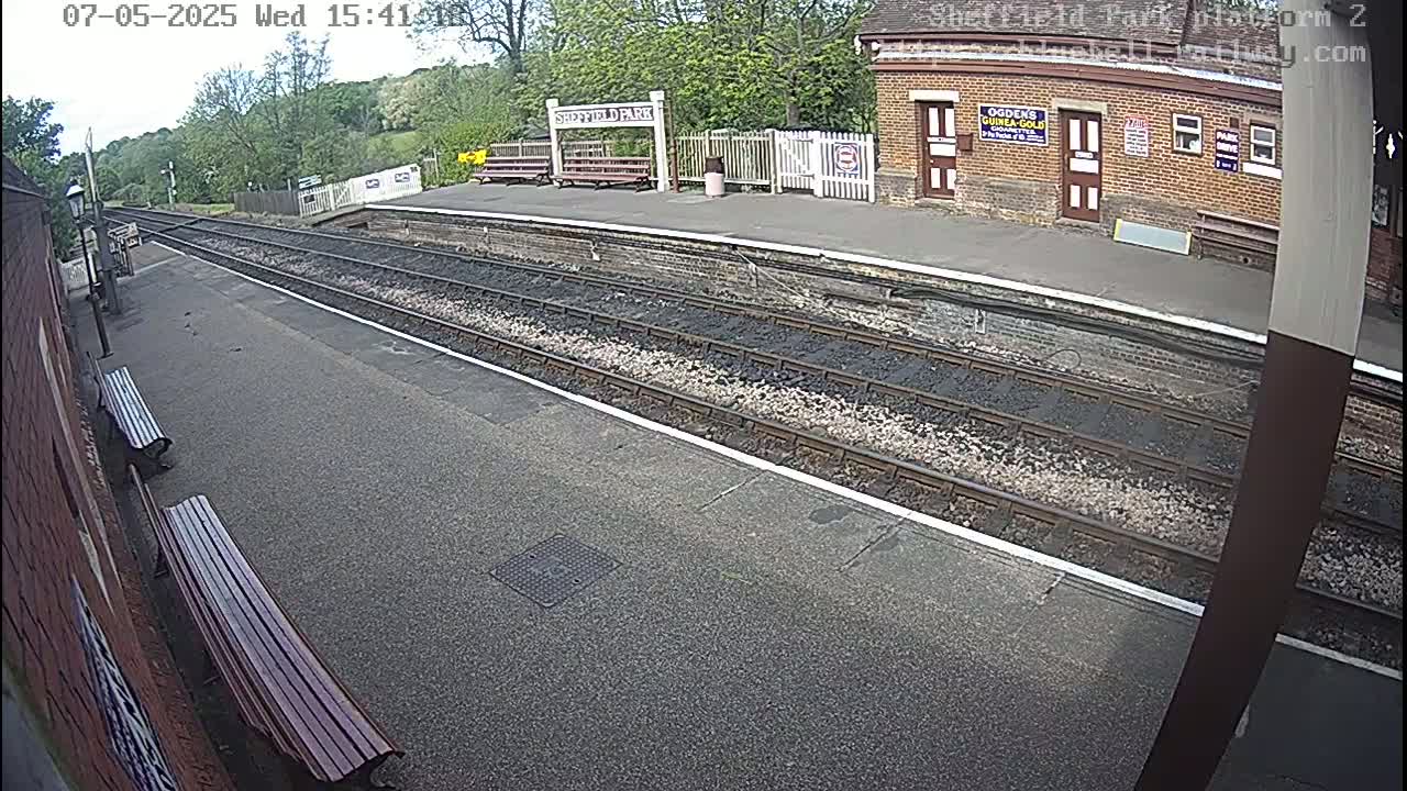 A mostly empty train platform on a sunny day, with train tracks extending into the distance and a brick building in the background.