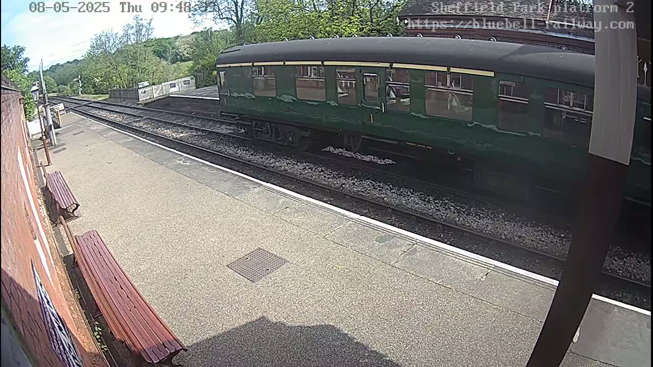 A dark-green passenger train car sits at a train station platform on a sunny day.