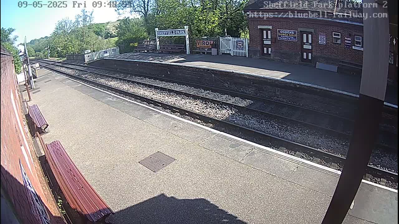 A sunny view of a mostly empty train platform with two wooden benches, train tracks, and a brick station building in the background.