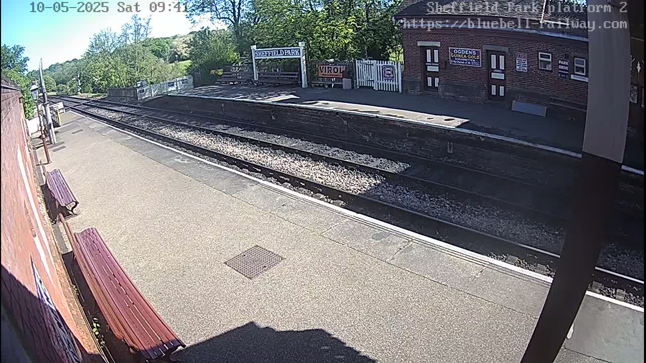 A sunny view of a train station platform with two benches, railway tracks, and a brick building in the background.