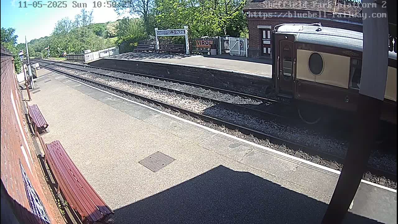 A railway carriage sits at a sunny train station platform next to two empty benches.