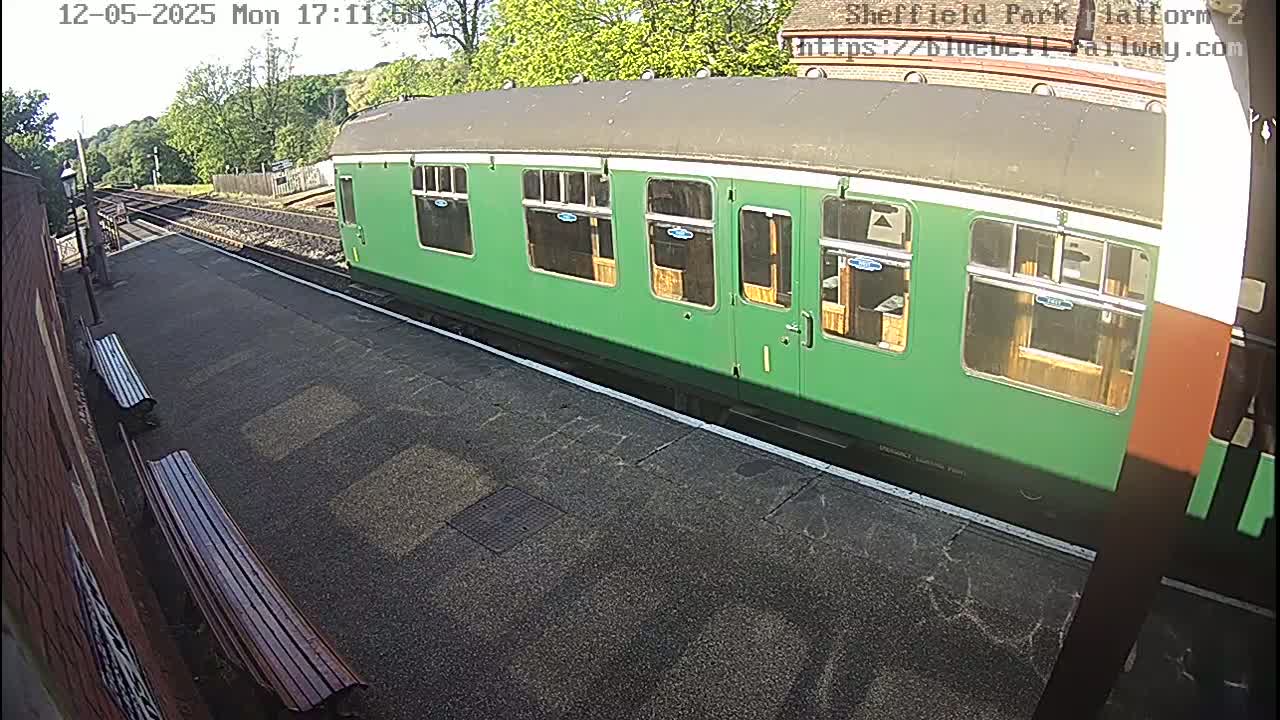 A long, green train car sits on a sunny train platform next to two wooden benches.