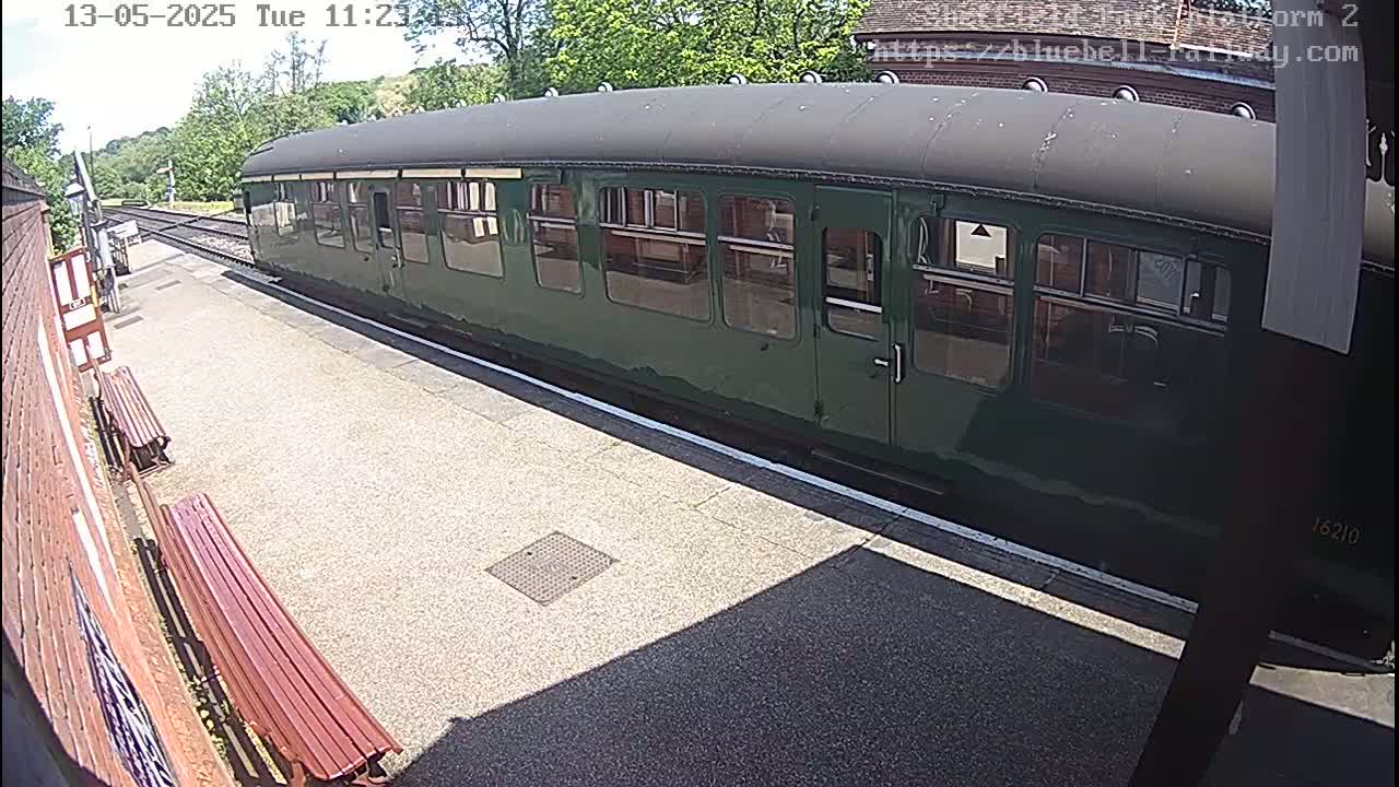 A dark-green railway carriage sits at a station platform on a sunny day.