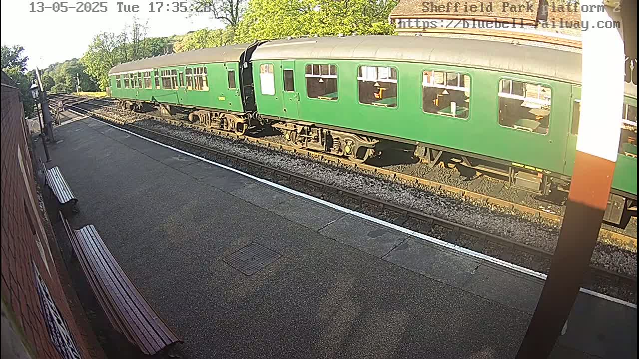 A long green train sits on a sunny railway platform next to two empty benches.