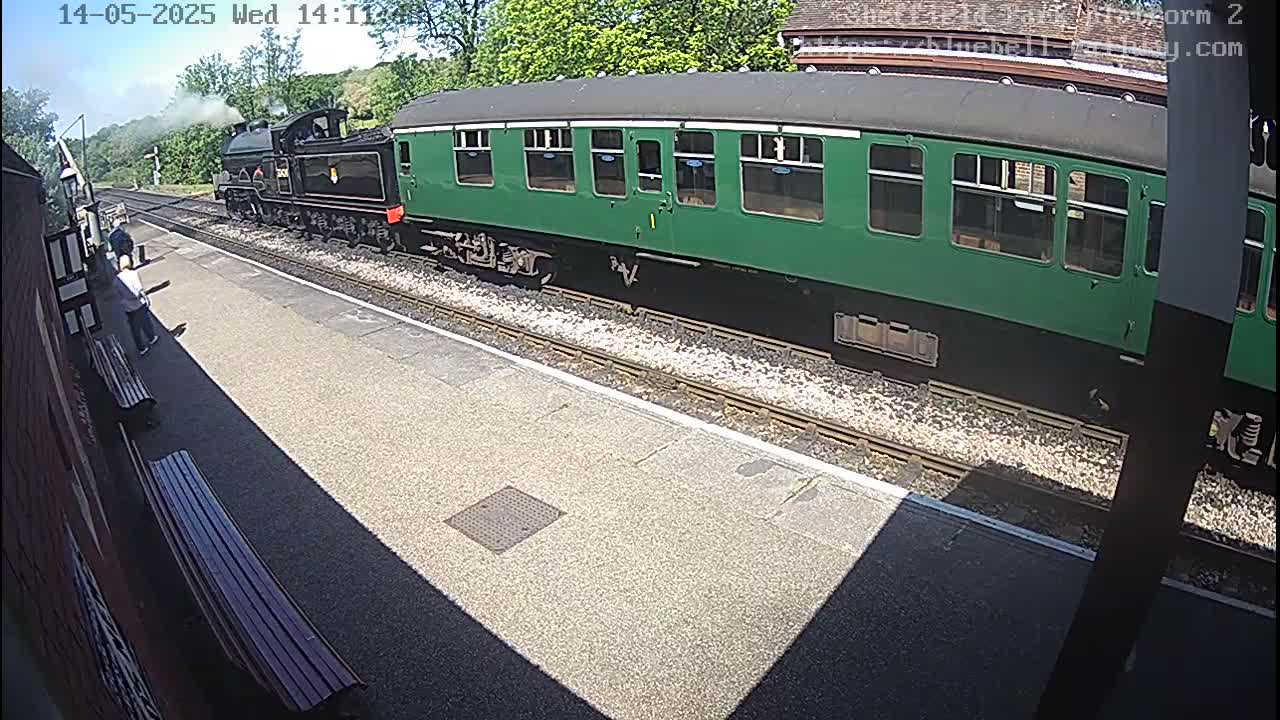 A dark-green passenger car is attached to a steam locomotive at a train station on a sunny day.