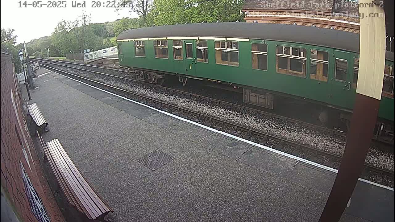 A long, dark-green passenger railcar sits on a train track at a station platform on an overcast day.