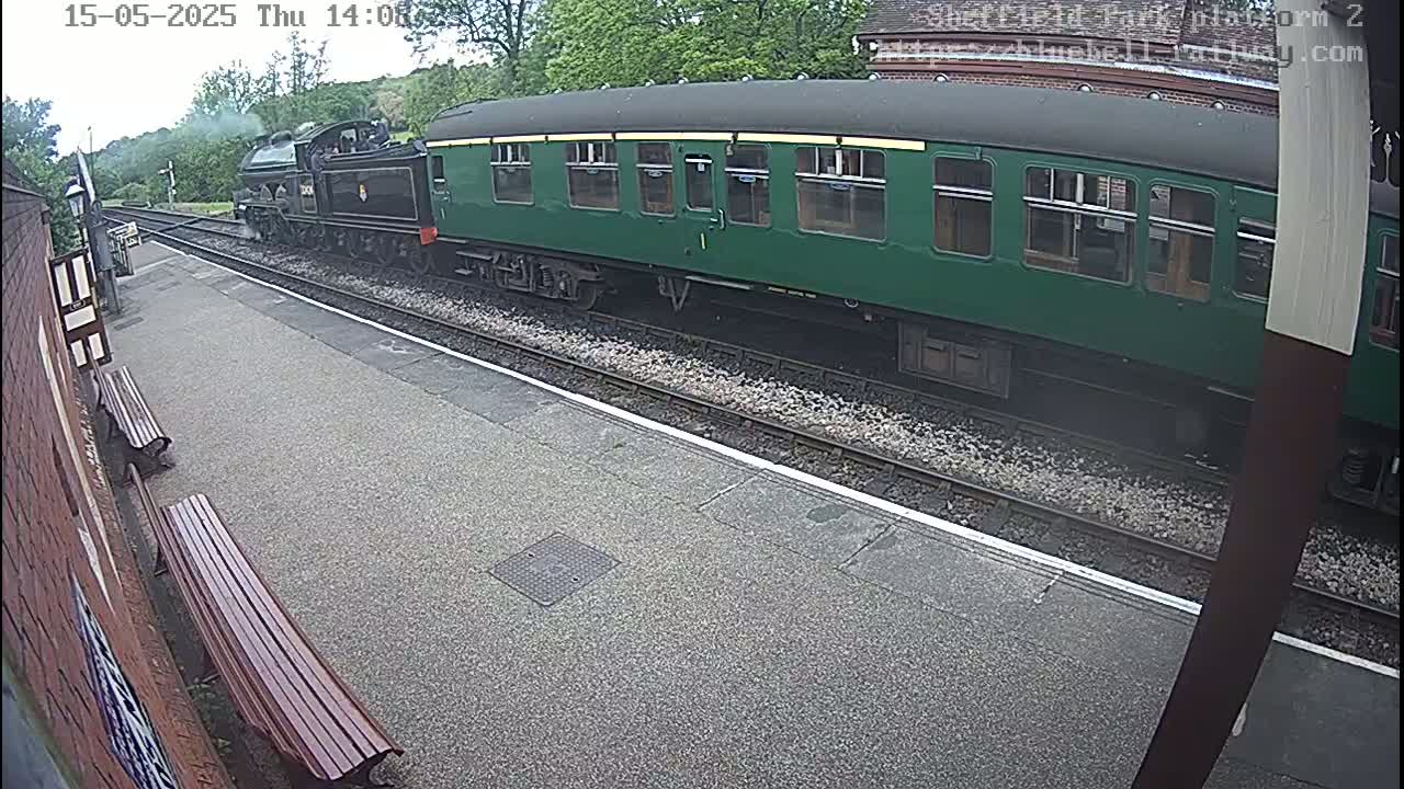 A dark-green passenger train car and a black steam locomotive are passing a train station platform on a partly cloudy day.
