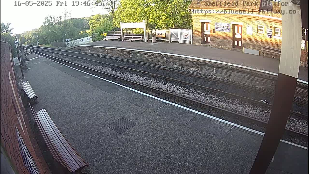 A mostly empty train platform with multiple train tracks and a brick building in the background on a partly sunny day.