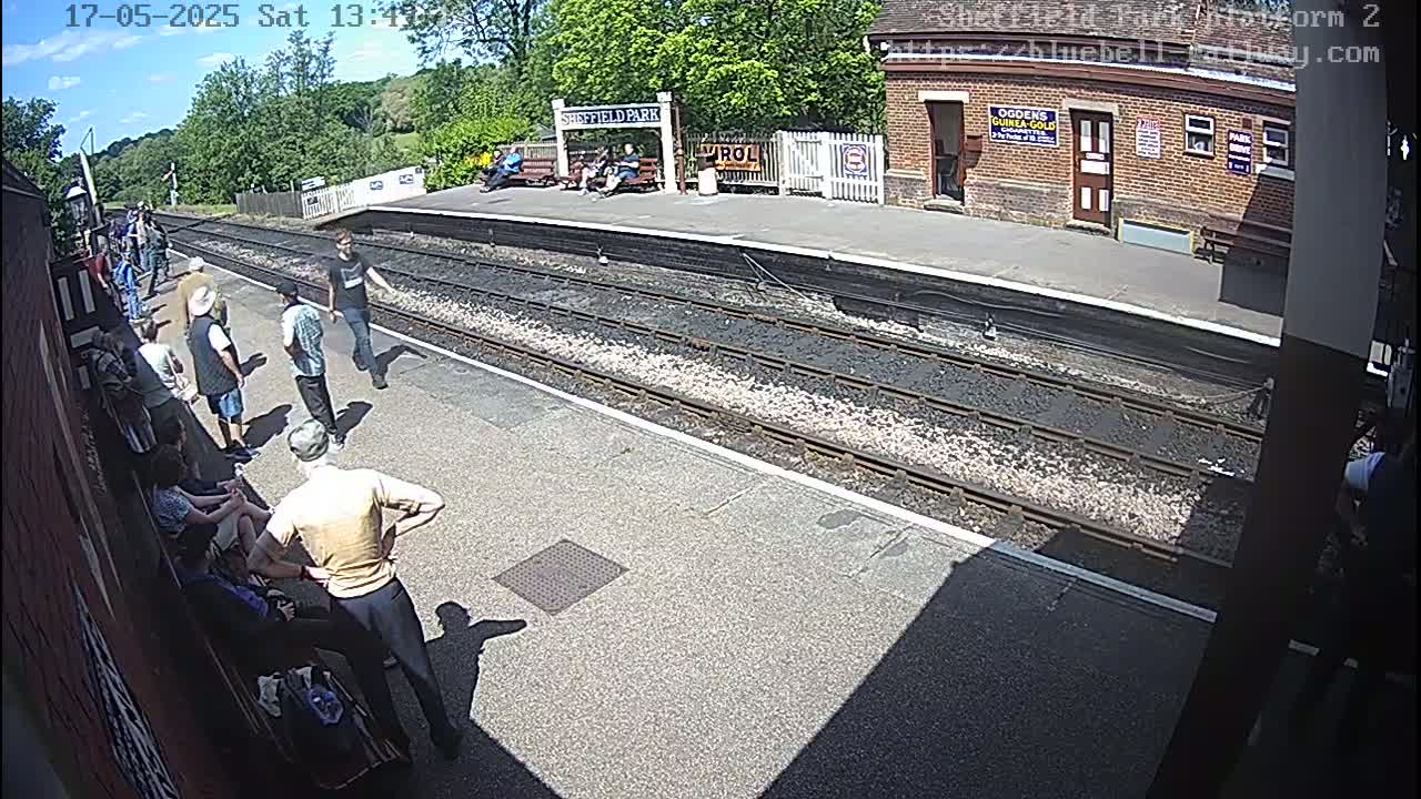 A sunny day at a train station platform shows several people standing and sitting on benches, with train tracks and a brick building in the background.