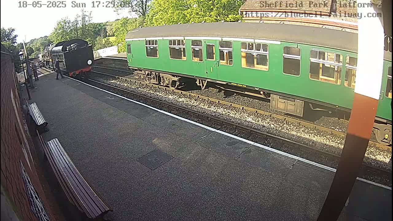 A steam locomotive and a long green passenger car are stopped at a train station on a sunny day.