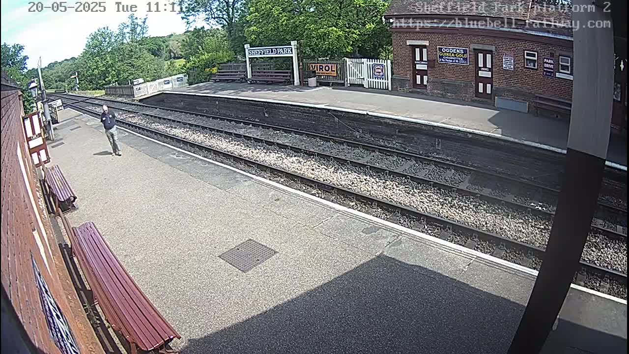 A sunny day at a train station platform shows a man walking on the platform with train tracks and a brick building visible in the background.