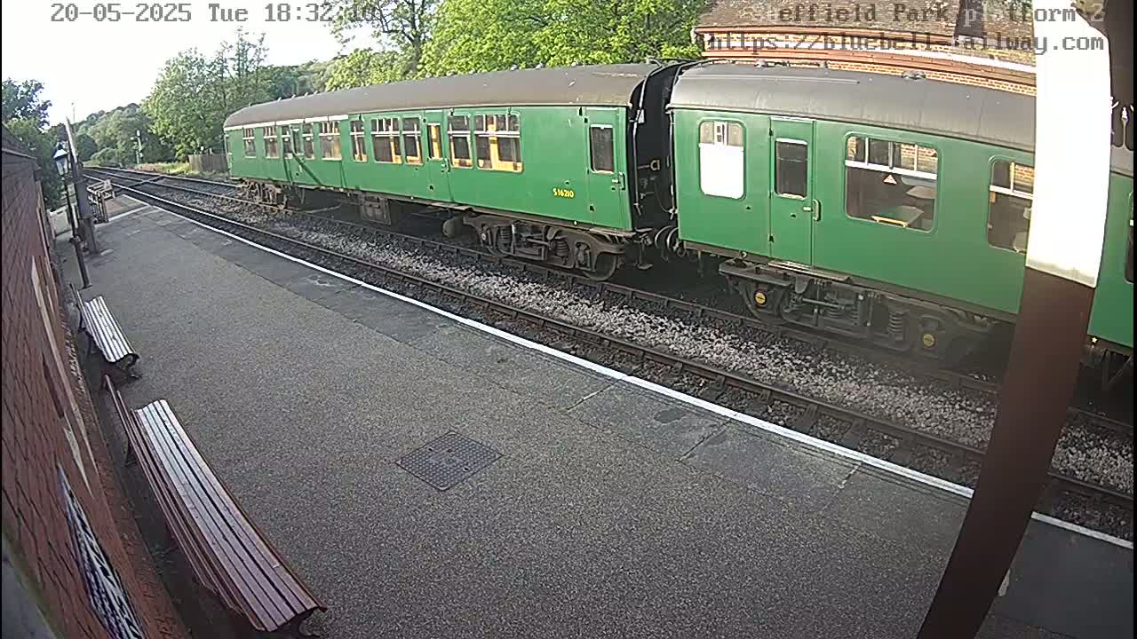 A long green train is stopped at a station platform on a partly sunny day.