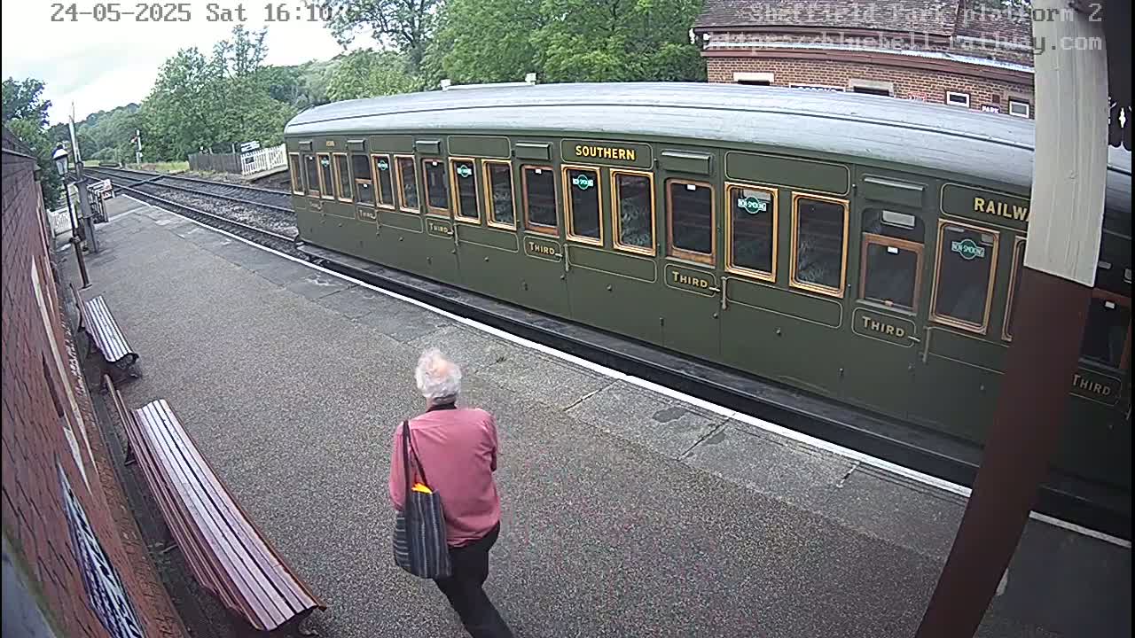 A person walks past a dark-green passenger train car at a train station on a partly sunny day.