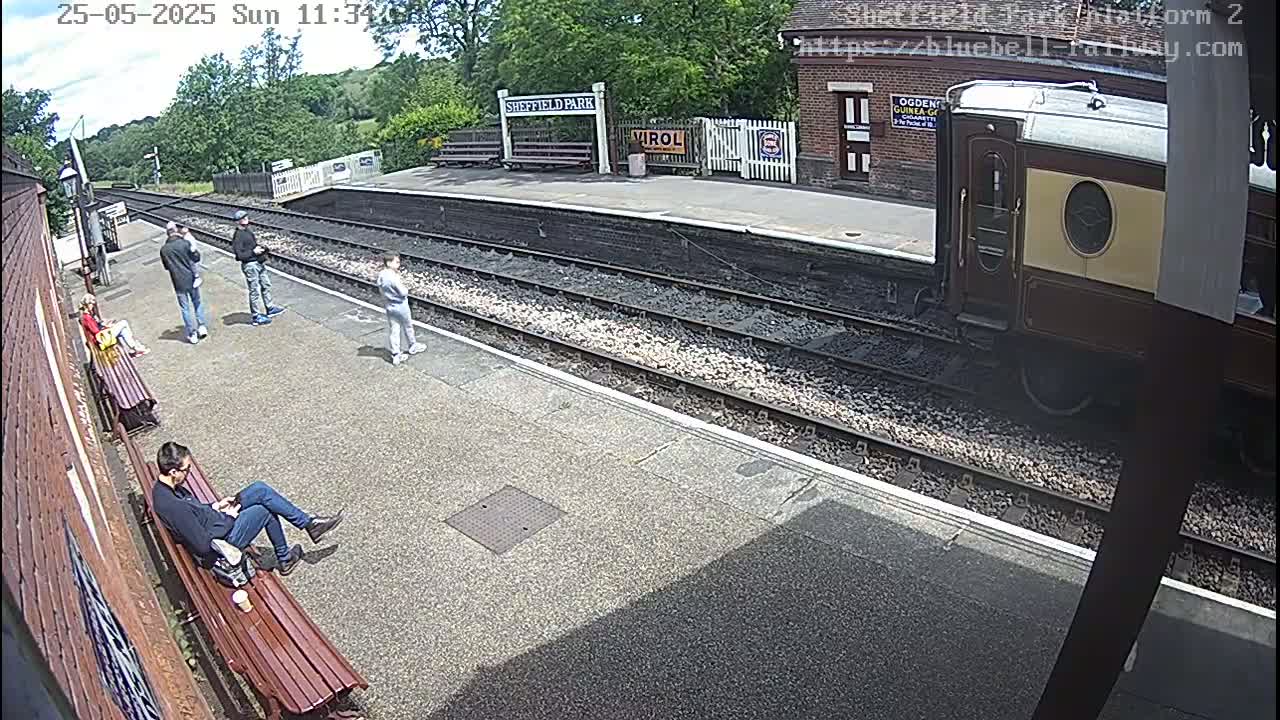 A sunny day at a train station shows several people standing and sitting on benches near train tracks, with a vintage train car partially visible.