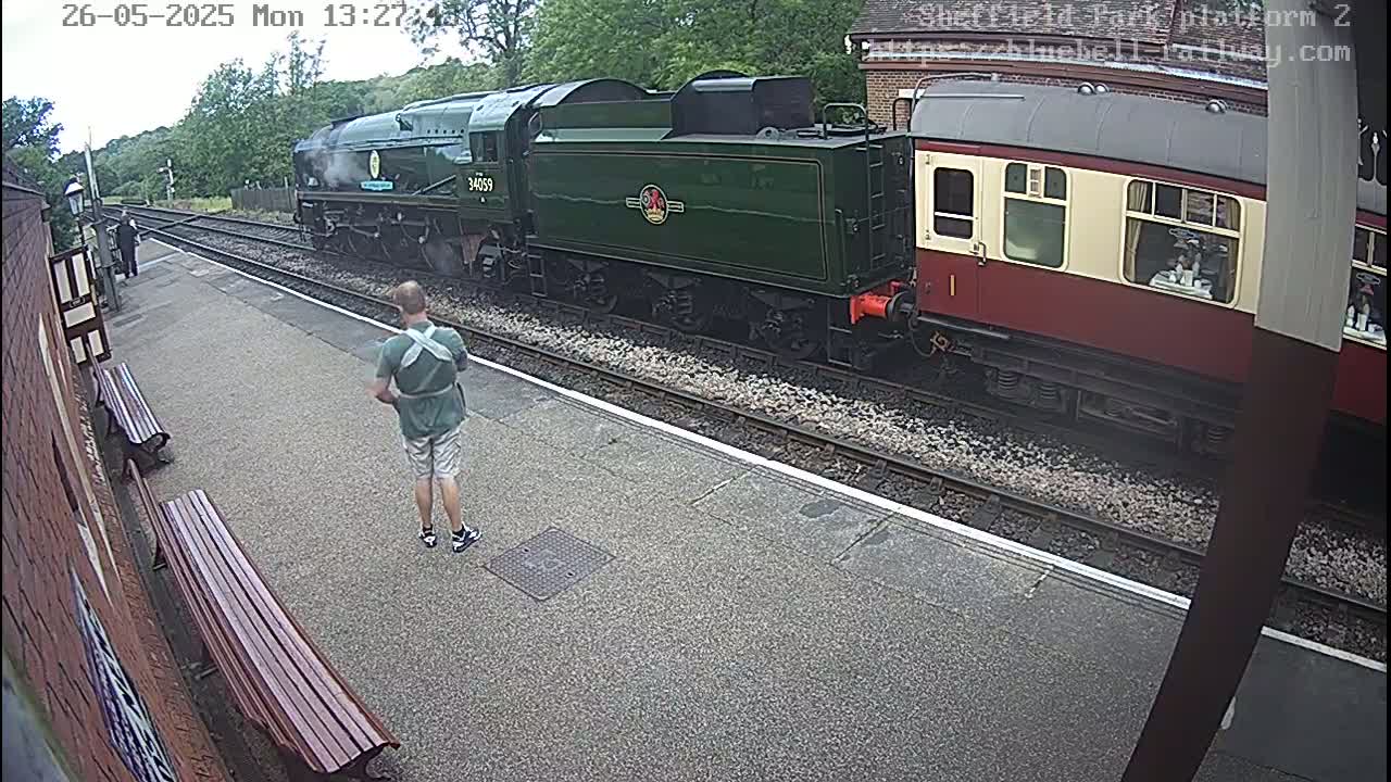 A green steam locomotive pulling a maroon passenger car sits at a train station platform on an overcast day.