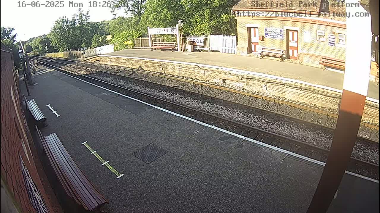 A sunny day at a train station platform shows train tracks, benches, and a brick station building.