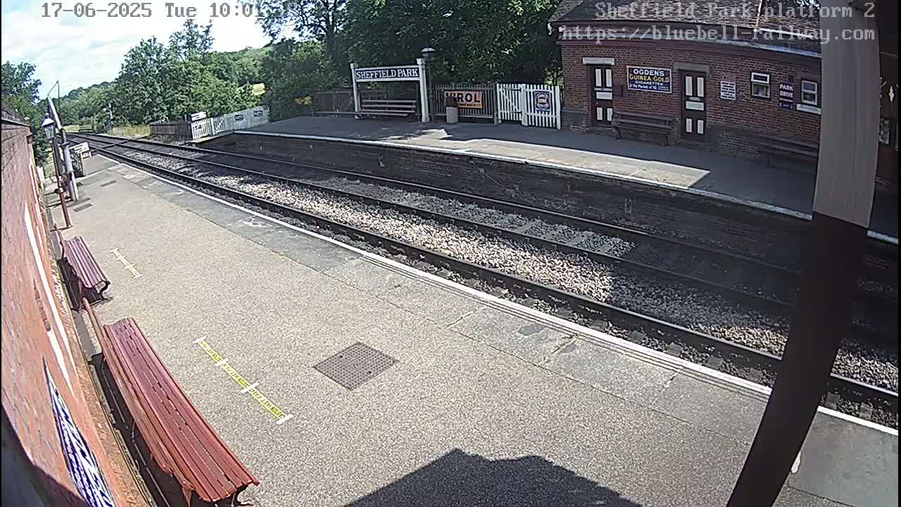 A sunny view of a train platform with two benches, train tracks, and a station building.
