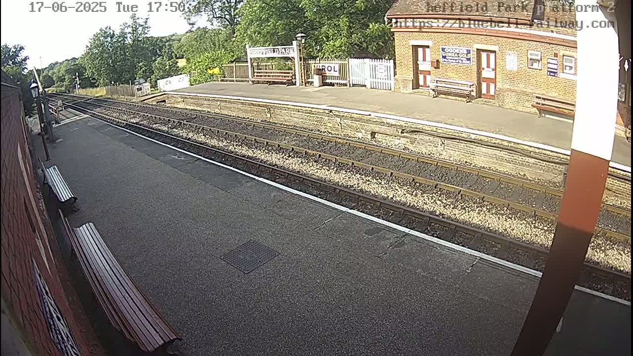A sunny view of a train platform with two benches, train tracks, and a brick building in the background.