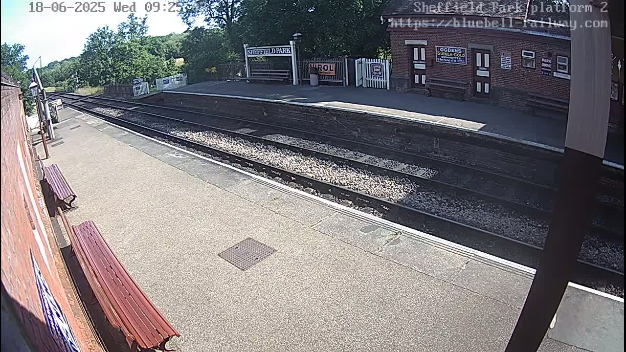 A sunny view of a train platform with two wooden benches, train tracks, and a small brick station building.
