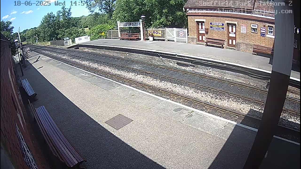 A sunny view of a train station platform with two parallel sets of train tracks, a brick building, and several benches.